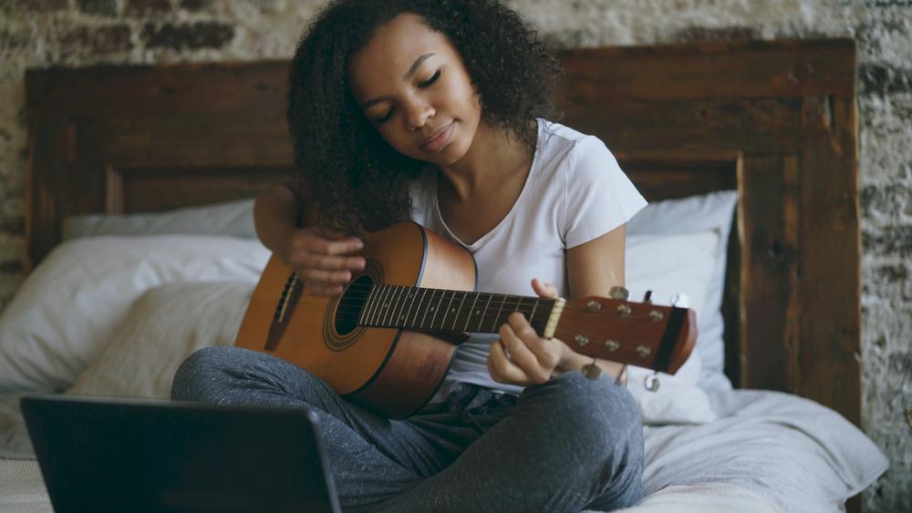 Attractive african american teenager girl concentraing learning to play acoustic guitar using laptop computer sitting on bed at home