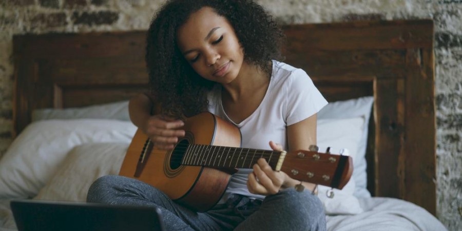 Attractive african american teenager girl concentraing learning to play acoustic guitar using laptop computer sitting on bed at home